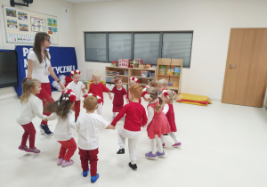 Dzieci i pani ubrani na biało - czerwono tańczą w kole, trzymając się za ręce.